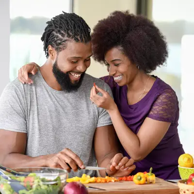 A smiling couple is preparing a meal together in a kitchen, surrounded by fresh vegetables.