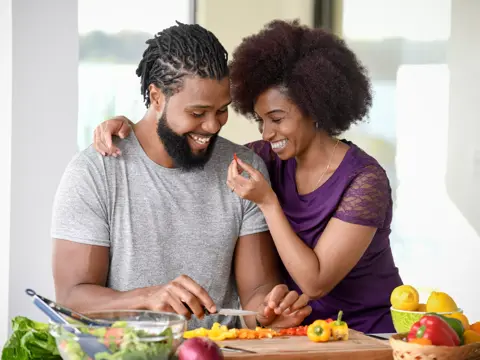 A smiling couple is preparing a meal together in a kitchen, surrounded by fresh vegetables.