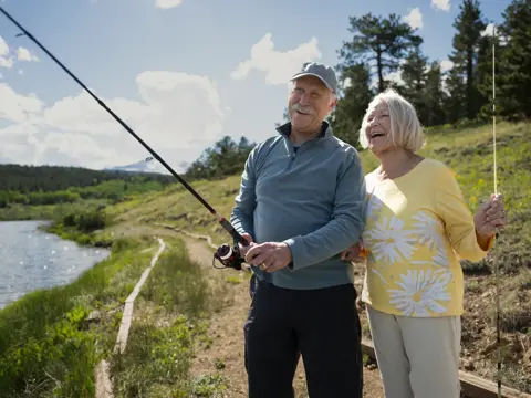 Elderly couple standing by a lake, holding fishing rods, smiling and enjoying their time together.