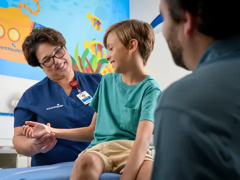 A nurse is examining a young boy's hand while his father watches, in a brightly decorated room.