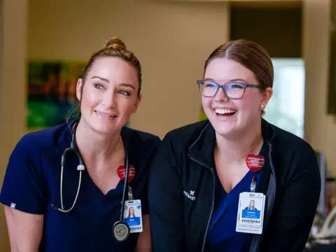 Two smiling nurses in blue uniforms stand together in a medical office, one wearing a stethoscope.