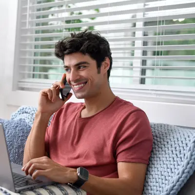 A man sitting on a couch with a laptop, smiling while talking on the phone.