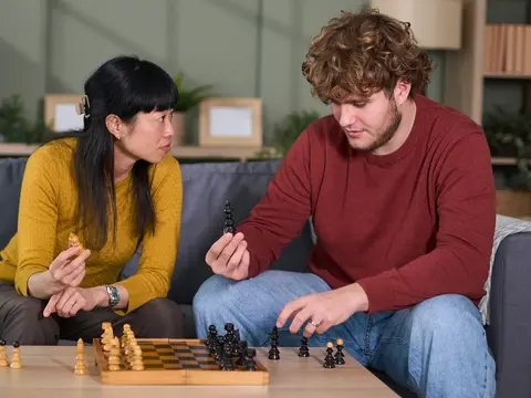 Man and woman sitting on a gray sofa playing chess together in a modern living room with plants and shelving in the background