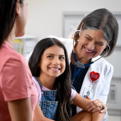 A smiling doctor with a stethoscope around her neck examines a young girl sitting on a bed, while a woman stands nearby.