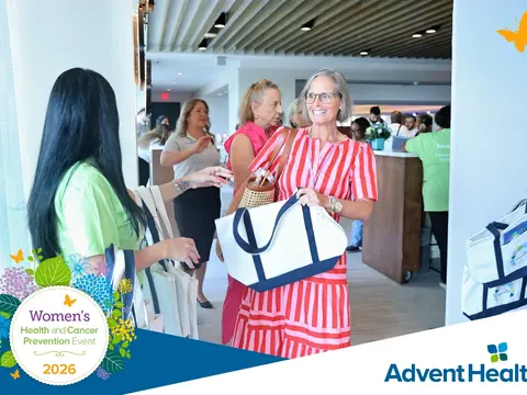 Women smiling and holding tote bags at the 2026 AdventHealth Women's Health and Cancer Prevention Event in a modern lobby with registration tables and attendees in the background