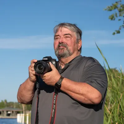 Photographer with gray beard holding a Canon DSLR camera outdoors near a waterfront with tall grasses and a clear blue sky