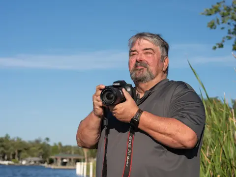 Photographer with gray beard holding a Canon DSLR camera outdoors near a waterfront with tall grasses and a clear blue sky