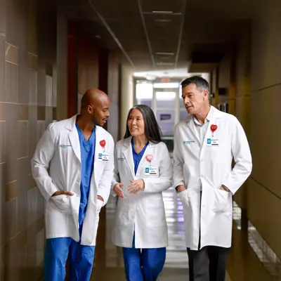 Three doctors in white coats and blue scrubs walk down a hospital hallway, smiling and conversing.