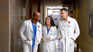 Three doctors in white coats and blue scrubs walk down a hospital hallway, smiling and conversing.