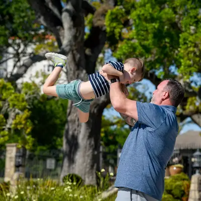 A man lifts a young boy high into the air in a park, both smiling.