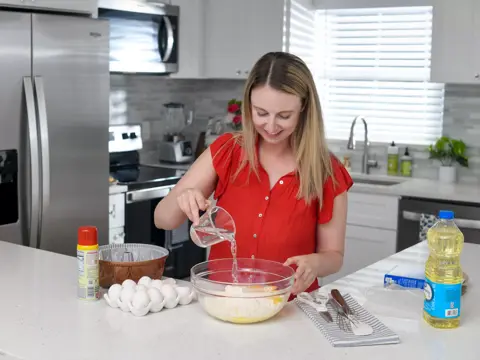 A woman in a red shirt pours water into a bowl of eggs and flour on a kitchen counter.