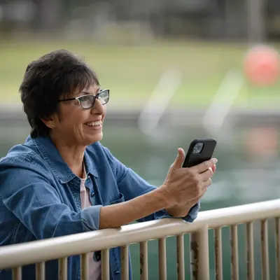 A smiling woman in a denim jacket stands by a railing, holding a smartphone.