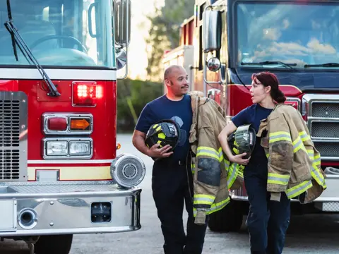 Two multiracial firefighters carrying helmets and fire protection suits, conversing as they walk between two fire engines.