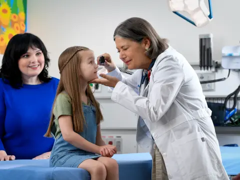 Doctor examining a young girl's eyes with a woman standing behind her in a medical setting.