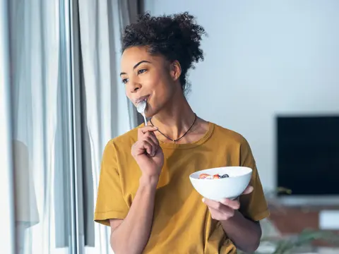 Woman in yellow shirt eating yogurt with berries from a white bowl while standing by a window in a bright home setting