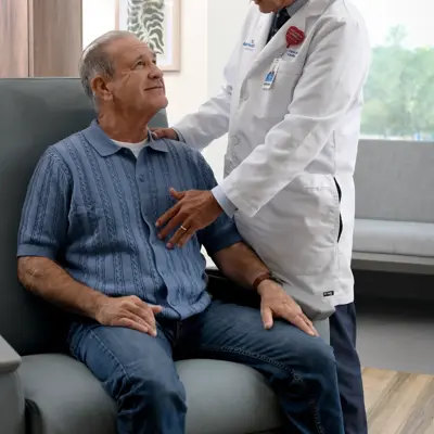 A doctor in a white coat examines a smiling elderly patient seated on a couch.