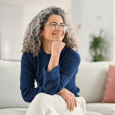 Smiling person with curly gray hair and glasses sits on a light sofa, resting their chin on one hand in a bright living room.