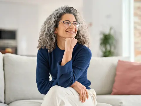 Smiling person with curly gray hair and glasses sits on a light sofa, resting their chin on one hand in a bright living room.