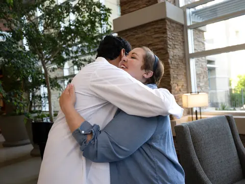 A smiling woman in a blue shirt embraces a man in a white coat in a bright, welcoming hospital lobby.