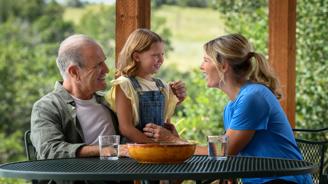 Grandfather, mother and daughter smiling and talking at a table in a garden.