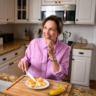 A woman in a pink shirt is cutting fruit on a cutting board in a kitchen.