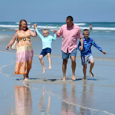 Family of four holding hands and walking on the beach, smiling and enjoying the day.