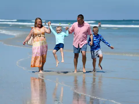 Family of four holding hands and walking on the beach, smiling and enjoying the day.