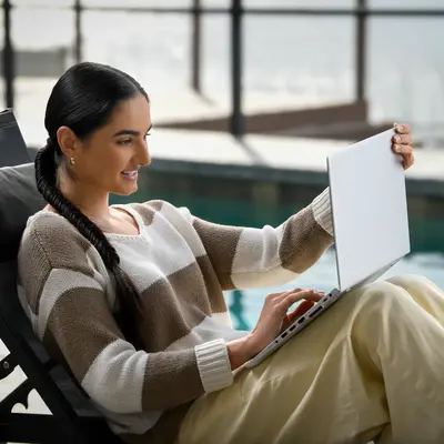 Hispanic woman with long braided hair sitting in a black lounge chair by an indoor pool, smiling while reviewing papers and using a laptop in a bright modern facility