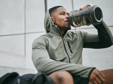 Man in gray hoodie drinking water from a large jug while sitting outdoors