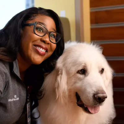 A smiling woman with glasses and an AdventHealth badge poses with a white dog.