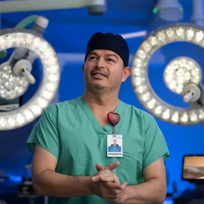 A surgeon in green scrubs stands in an operating room with surgical lights overhead.