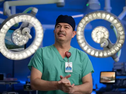 A surgeon in green scrubs stands in an operating room with surgical lights overhead.