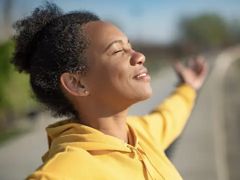 A woman standing in the sun, smiling, with her eyes closed.