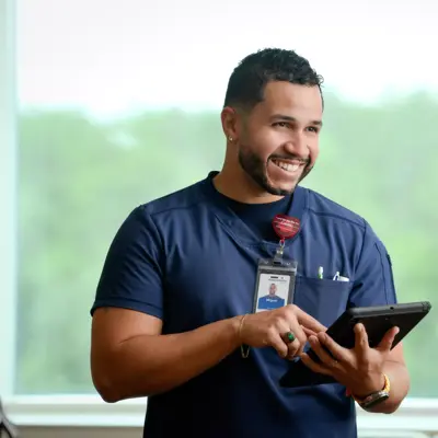A smiling healthcare worker in scrubs holds a tablet, standing by a window with trees outside.