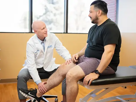 A physician in a white coat examines a patient's knee during an orthopedic consultation in a clinical exam room
