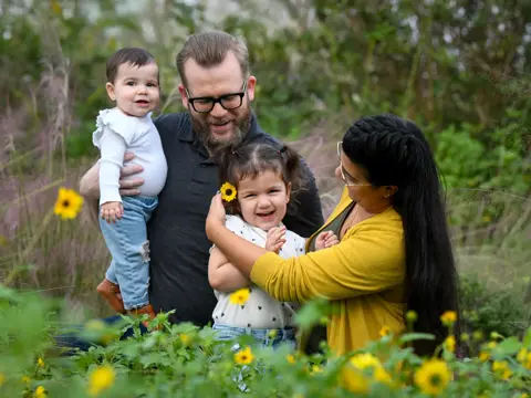 A family of four, consisting of a man, a woman, and two children, smiling in a field of flowers.
