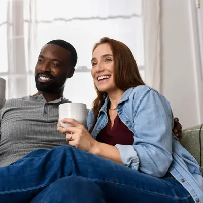 Smiling couple sitting together on a couch holding coffee mugs in a bright living room with natural light