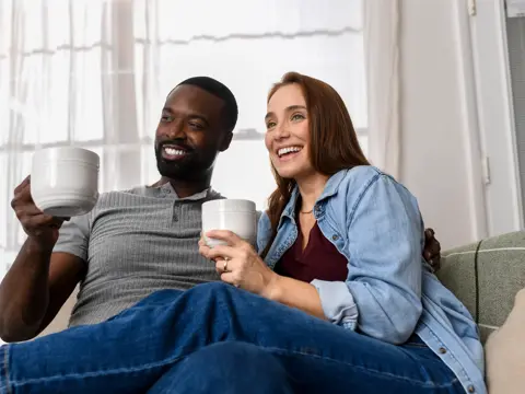 Smiling couple sitting together on a couch holding coffee mugs in a bright living room with natural light