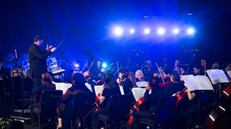 Conductor leading an orchestra on stage with blue lighting, musicians playing various instruments.