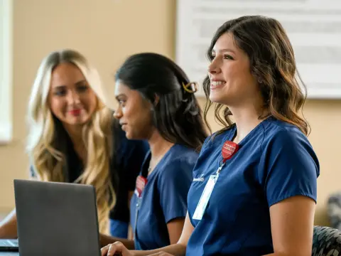 Three smiling nurses in blue uniforms work on laptops in a room.