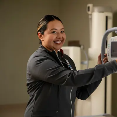 A smiling woman in a black jacket holds a medical imaging device in a hospital room.
