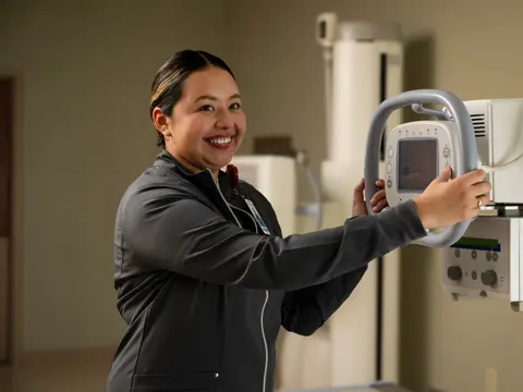 A smiling woman in a black jacket holds a medical imaging device in a hospital room.