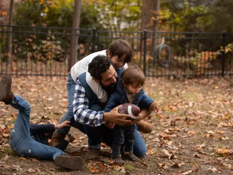 A father and two children are playing with a football in a park, surrounded by fallen leaves.