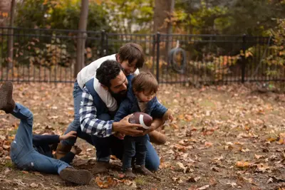 A father and two children are playing with a football in a park, surrounded by fallen leaves.