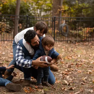 A father and two children are playing with a football in a park, surrounded by fallen leaves.