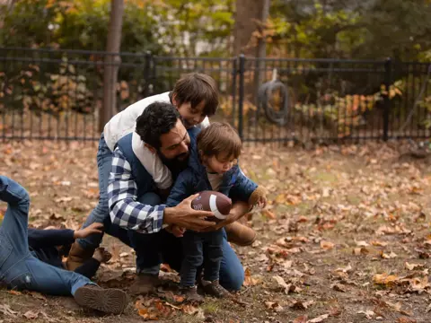 A father and two children are playing with a football in a park, surrounded by fallen leaves.