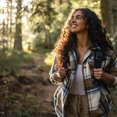 Smiling person with curly hair wearing a plaid jacket and backpack walking on a forest trail.