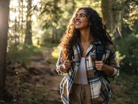 Smiling person with curly hair wearing a plaid jacket and backpack walking on a forest trail.