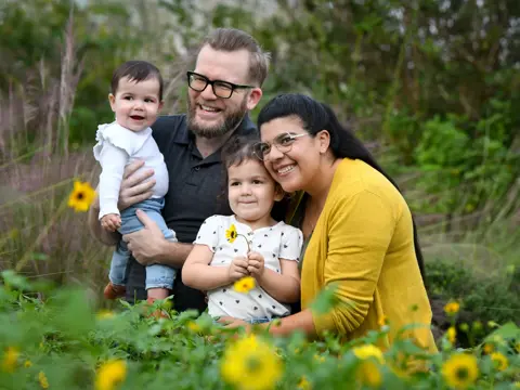 A family of four, two adults and two children, smiling and posing in a field of flowers.