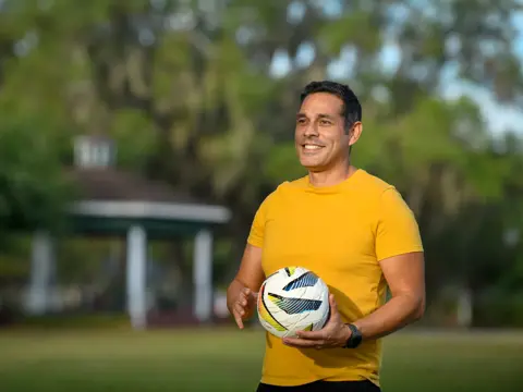 A man in a yellow shirt holds a soccer ball in a park.